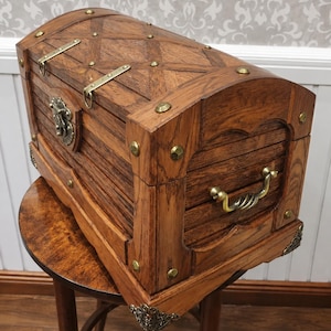 May include: A rectangular wooden chest with a curved lid, featuring a dark brown wood grain finish. The chest is accented with brass-coloured metal corner plates, decorative studs, two top latches, a front locking mechanism, and a side handle.