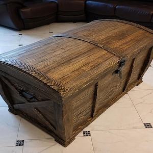 May include: A rectangular wooden storage chest with a dark brown stained finish and a curved, barrel-style lid. The chest features visible wood grain, black metal side handles, and a central black metal latch mechanism on the front panel.