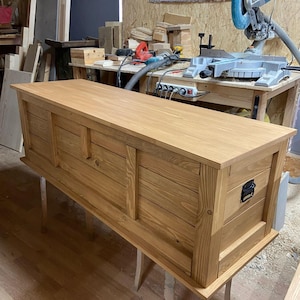 May include: A rectangular wooden storage chest with a natural light brown finish, featuring horizontal plank paneling on the front and sides, a flat top surface, and a black metal handle mounted on the side panel for lifting and carrying.