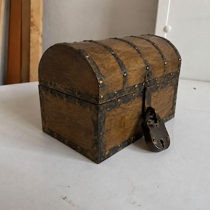 May include: A small, wooden treasure chest with a curved top and metal accents. The chest is brown with dark metal bands and a matching lock. The chest is closed and ready to store valuables.