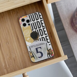 May include: A smartphone case featuring a graphic of a footballer wearing a white jersey with the name BELLINGHAM and the number 5 on the back. The design includes the repeated text JUDE in black and a gold-toned section with the text MADE IN SPAIN.