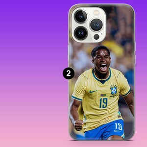 May include: A clear smartphone case featuring a printed image of a soccer player wearing a yellow Brazil jersey with the number 19 in blue, blue shorts, and a celebratory expression. The case is shown on a phone with a triple-lens camera system.