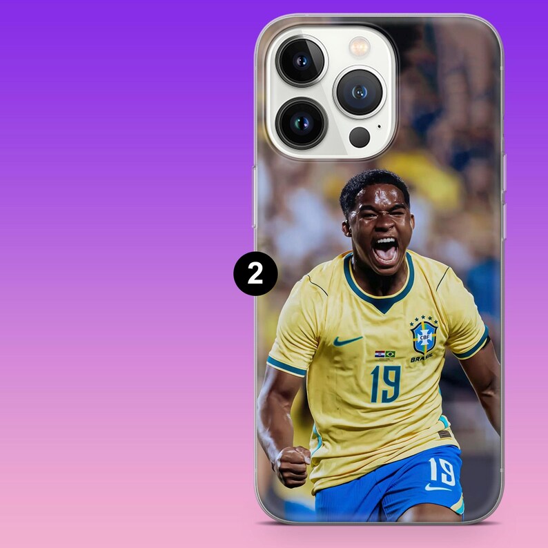 May include: A clear smartphone case featuring a printed image of a soccer player wearing a yellow Brazil jersey with the number 19 in blue, blue shorts, and a celebratory expression. The case is shown on a phone with a triple-lens camera system.