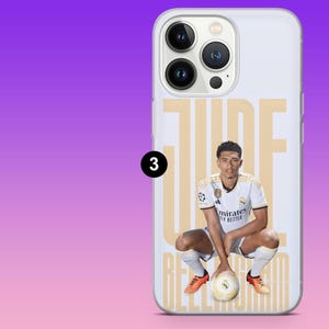 May include: A clear smartphone case featuring a graphic of a football player in a white kit crouching with a football. The name JUDE BELLINGHAM is printed in large tan letters behind the player. The case has a cutout for a triple camera system.