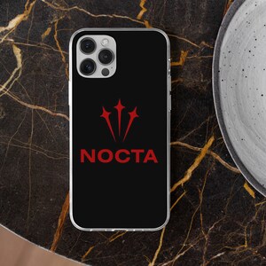 K&ouml;nnte beinhalten: Schwarze Handyh&uuml;lle mit rotem Logo und dem Wort "NOCTA". Die H&uuml;lle hat einen transparenten Rand und einen Kameraausschnitt. Das Logo ist ein stilisiertes Dreizack-Design. Die Handyh&uuml;lle liegt auf einer dunklen, gemusterten Oberfl&auml;che.