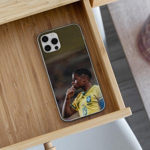 May include: A smartphone case featuring a printed image of a soccer player wearing a yellow Brazil national team jersey with the number 19 visible. The player is shown in profile, touching their lips with their fingers in a celebratory gesture.