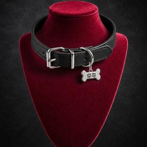 Może przedstawiać: Czarna obroża z metalową klamrą w kolorze srebrnym oraz zawieszką w kształcie kości z wyciętym wzorem łapy. Obroża ma fakturowaną powierzchnię i metalowe kółko w kształcie litery D do przymocowania wiszącej zawieszki.