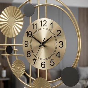 Puede incluir: Un reloj de pared con marco circular dorado y barras verticales, una esfera central dorada con números negros del 1 al 12 y manecillas negras. El diseño incluye detalles dorados en forma de abanico plisado y discos negros sólidos.