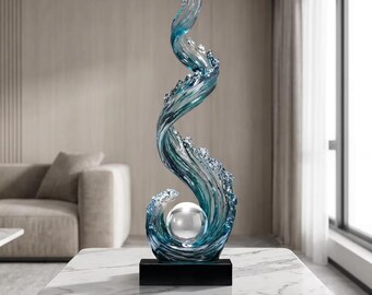 May include: A tall, abstract sculpture with a swirling, wave-like design. The sculpture is primarily blue and clear, with a silver sphere at its center. It sits on a black rectangular base, placed on a white marble table. The artwork evokes a sense of movement and fluidity.