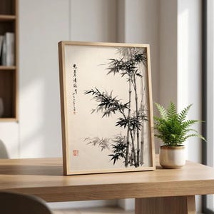 Puede incluir: Una lámina de arte enmarcada con una pintura a tinta negra de tallos y hojas de bambú sobre un fondo color crema. La obra incluye caracteres de caligrafía asiática en negro vertical y un pequeño sello cuadrado de color rojo en la esquina.