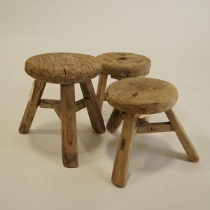 May include: Three small, round stools crafted from light brown, weathered wood. Each stool features a circular seat with a visible grain pattern and three sturdy, angled legs connected by a triangular support structure beneath the seat.