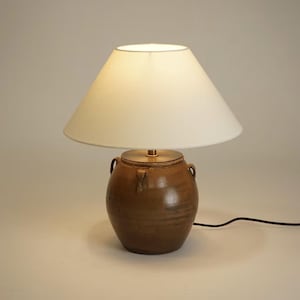 May include: A table lamp featuring a rounded brown ceramic base with two small side handles and a smooth, tapered off-white fabric lampshade. The lamp includes a black power cord extending from the base, which sits on a flat surface.