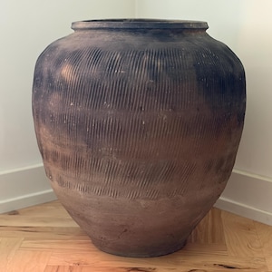 Peut inclure: Une grande jarre en céramique brun foncé, de forme arrondie, dotée d'une large ouverture et d'un bord épais. La surface extérieure présente un motif texturé de rainures verticales sculptées sur les parties supérieure et médiane.