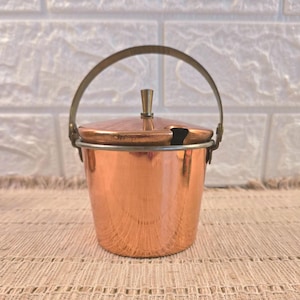 May include: A polished copper bucket with a matching lid and a thin metal handle. The lid features a small central knob and a cutout notch on the side. The bucket has a tapered cylindrical shape with a smooth, reflective metallic finish.
