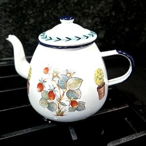 May include: A white enamel teapot featuring a painted strawberry plant with red berries and green leaves, flanked by two small potted yellow topiary trees. The lid has a light blue leaf border, and the handle, spout, and lid rim are dark blue.