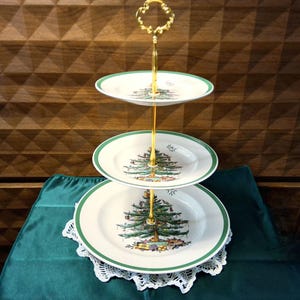 May include: A three-tiered serving stand featuring white ceramic plates with a green rim and a central Christmas tree illustration. The stand has a gold-colored metal central rod and a decorative handle at the top for easy transport.