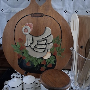 May include: A round wooden cutting board with a heart-shaped handle cutout, featuring a hand-painted gray chicken sitting in a black pot surrounded by red strawberries. Beside it are white ceramic salt and pepper shakers and a lidded jar.