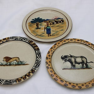May include: Three round ceramic plates with beige centers and decorative rims. One plate features a landscape with a figure, one shows a lion in grass with a black and white geometric border, and one depicts a buffalo with a spotted brown border.