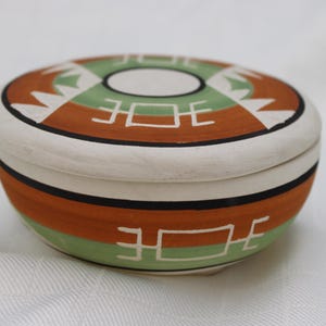 May include: A round ceramic lidded box featuring a hand-painted design with bands of terracotta orange, sage green, and white. The lid and base are decorated with white geometric symbols and thin black lines encircling the circumference of the vessel.