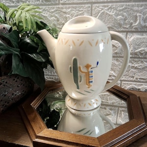 May include: A cream-colored ceramic teapot featuring a lid and a handle. The body is decorated with a painted green cactus and a tan figure wearing a blue and white striped garment. A pattern of small tan leaf-like shapes encircles the top and base.