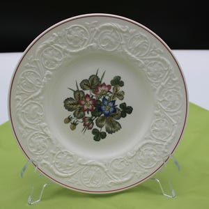 May include: A round white ceramic plate featuring a central botanical illustration of green leaves, small red strawberries, and blue and pink flowers. The rim is decorated with an embossed scrollwork pattern and a thin red line along the outer edge.