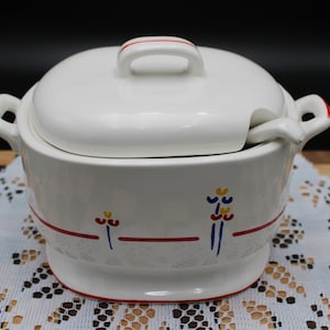 May include: A white ceramic tureen with a matching lid and a small ladle. The tureen features a horizontal red line accented with small blue and yellow floral motifs. It has two side handles and a raised base, with red trim on the lid handle.
