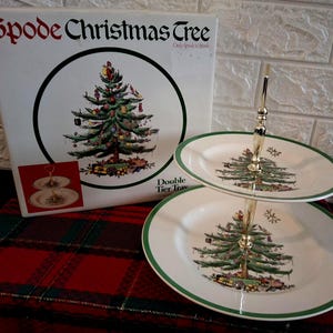 May include: A Spode Christmas Tree double tier tray featuring two white ceramic plates with green rims and a central Christmas tree illustration. The set includes a gold-tone metal handle and a white cardboard box with the text Spode Christmas Tree.