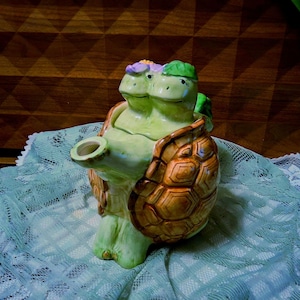 May include: A ceramic teapot shaped like a turtle with a light green body and a brown, textured shell. The lid features two small turtle heads, one adorned with a purple flower and the other with a green leaf. The spout extends from the front.