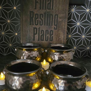 May include: Four metallic, hammered-finish cauldrons arranged in a square formation. Behind them stands a rectangular wooden sign with the text "Final Resting Place" engraved in black. Small, white LED tea lights are placed between the cauldrons.