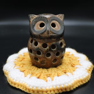 May include: A dark brown ceramic owl figurine with large circular eyes and a body featuring multiple small, round cut-out holes. The owl is positioned on a round, crocheted coaster with a yellow center and a white scalloped border edge.