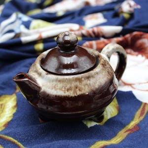 May include: A small, brown ceramic teapot with a lid and handle. The teapot has a rounded shape and sits on three small feet. The exterior is a glossy brown with a lighter, speckled band around the middle. The teapot is set against a blue floral background.