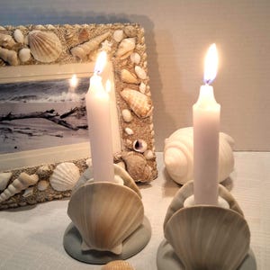 May include: Two white taper candles burning in beige scallop shell candle holders with gray circular bases. A rectangular picture frame decorated with various seashells and a black and white beach photograph sits behind the candles.