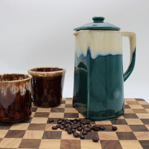 May include: A ceramic coffee set consisting of a tall, square-based pitcher with a dark teal and cream drip glaze, and two matching brown mugs with a cream drip glaze. A small pile of roasted coffee beans sits between the pitcher and the mugs.