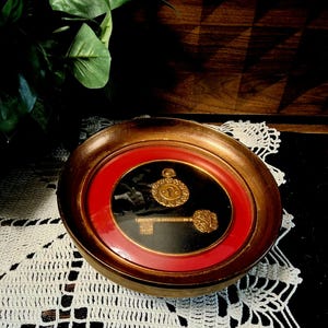 May include: A circular wall plaque featuring a gold-toned key and a circular emblem with a keyhole design centered on a black background. The piece is framed by a vibrant red inner border and a wider, distressed brown wooden outer frame.