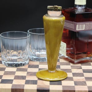 May include: A tall, olive-green ceramic table lighter with a metallic top mechanism, positioned alongside two clear glass tumblers and a square glass bottle containing a dark amber liquid with a visible white label and barcode.