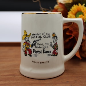 May include: A cream-colored ceramic beer mug with a gold-trimmed rim and handle. The front features a cartoon graphic of two figures, the text Member of the PISTOL CLUB, Drink Till Midnight, Pistol Dawn, I.P. FREELY PRES, P. WEE SECY, and SOUTH DAKOTA.