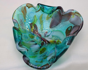 Peut inclure: Un bol décoratif en verre avec un bord ondulé et organique. Le bol présente une couleur de base turquoise et sarcelle vibrante, accentuée par des motifs tourbillonnants de blanc, de violet et de flocons dorés incrustés dans le verre.