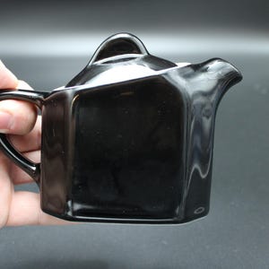 May include: A small, glossy black ceramic teapot with a distinct geometric, angular body shape. It features a curved handle on the left side, a short spout on the right, and a matching black lid that sits flush with the top of the vessel.