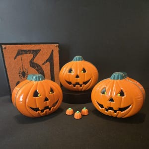 May include: Three orange jack-o'-lantern containers with green stems and carved faces, accompanied by three miniature orange pumpkin figurines. Behind them stands a square orange sign featuring the black number 31 and a spider illustration.