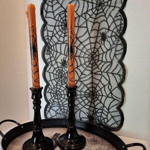 May include: Two orange taper candles featuring a black spiderweb print, each held in a glossy black candlestick holder. These are placed on a black metal tray with a graphic print, set against a black lace runner with a spiderweb pattern.