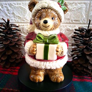 May include: A ceramic bear figurine dressed in a red and white Santa suit with a matching hat. The bear has brown fur and is holding a white gift box tied with a green ribbon. A small sprig of green holly with red berries is attached to the hat.