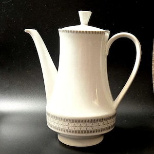 May include: A white porcelain coffee pot featuring a tapered body, a curved handle, and a long spout. The lid and the base are decorated with a repeating grey geometric pattern consisting of small triangles and star-like shapes in a band.