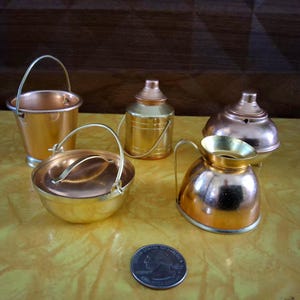 May include: A collection of four miniature metal kitchen vessels in copper and gold tones, including a small bucket with a handle, a lidded pot, a canister, and a pitcher, displayed alongside a standard United States quarter coin for scale.