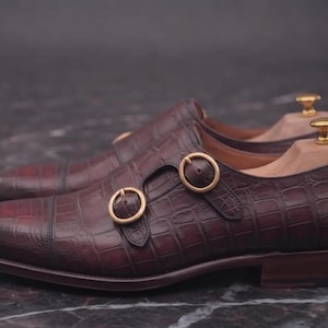 May include: A pair of dark burgundy double monk strap shoes featuring a textured crocodile-embossed leather finish, rounded toes, and two gold-toned circular buckles on each shoe. Each shoe includes a light-coloured wooden shoe tree insert.