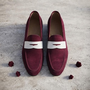 May include: A pair of deep burgundy suede loafers featuring a contrasting white leather saddle strap across the vamp. The shoes have a rounded toe design and a smooth interior lining, presented with small dark red decorative floral accents.