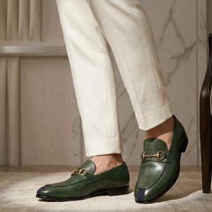 May include: A pair of olive green leather loafers featuring a gold-toned metal horsebit detail across the vamp and a dark blue vertical stripe accent on the rounded toe. The shoes have a low heel and a smooth finish, paired with white trousers.