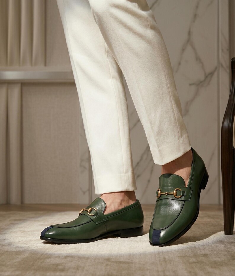May include: A pair of olive green leather loafers featuring a gold-toned metal horsebit detail across the vamp and a dark blue vertical stripe accent on the rounded toe. The shoes have a low heel and a smooth finish, paired with white trousers.