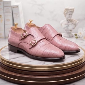 May include: A pair of light pink double monk strap shoes featuring a textured crocodile skin pattern, gold-toned metal buckles, and a dark brown sole. Each shoe includes a wooden shoe tree inserted inside to maintain the shape of the footwear.