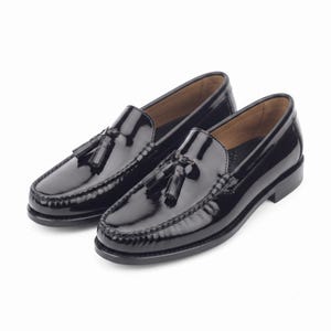 May include: A pair of black patent leather loafers featuring a classic moccasin-style toe construction, a low stacked heel, and decorative tassels on the vamp. The shoes have a polished, high-gloss finish and a smooth, rounded silhouette.