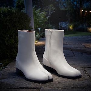 May include: A pair of white leather ankle boots with a split-toe design. The boots have a side zipper and a low, black heel. The boots are set on a stone patio with a blurred background of greenery and a white column.
